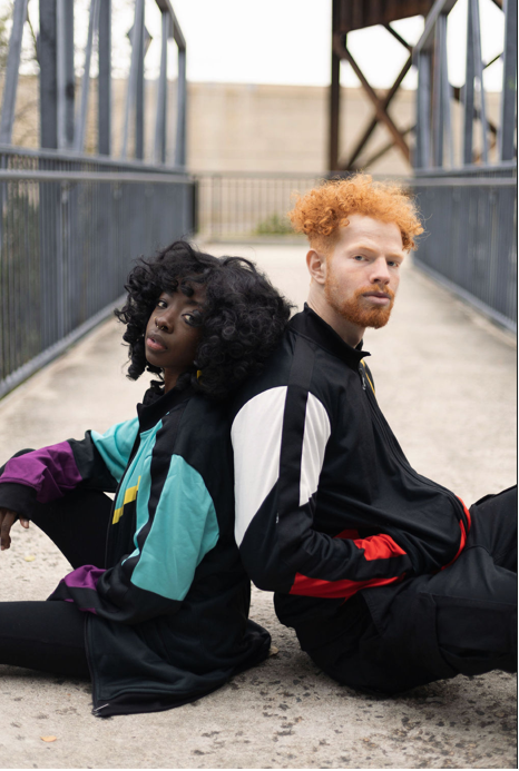 Two people sitting on a concrete surface wearing colorful tracksuits.